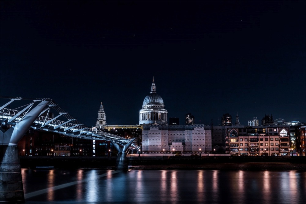 A dramatic photograph taken at night time showing the Millennium Bridge leading to the Thames riverside and buildings in front of St Paul's Cathedral Dome.